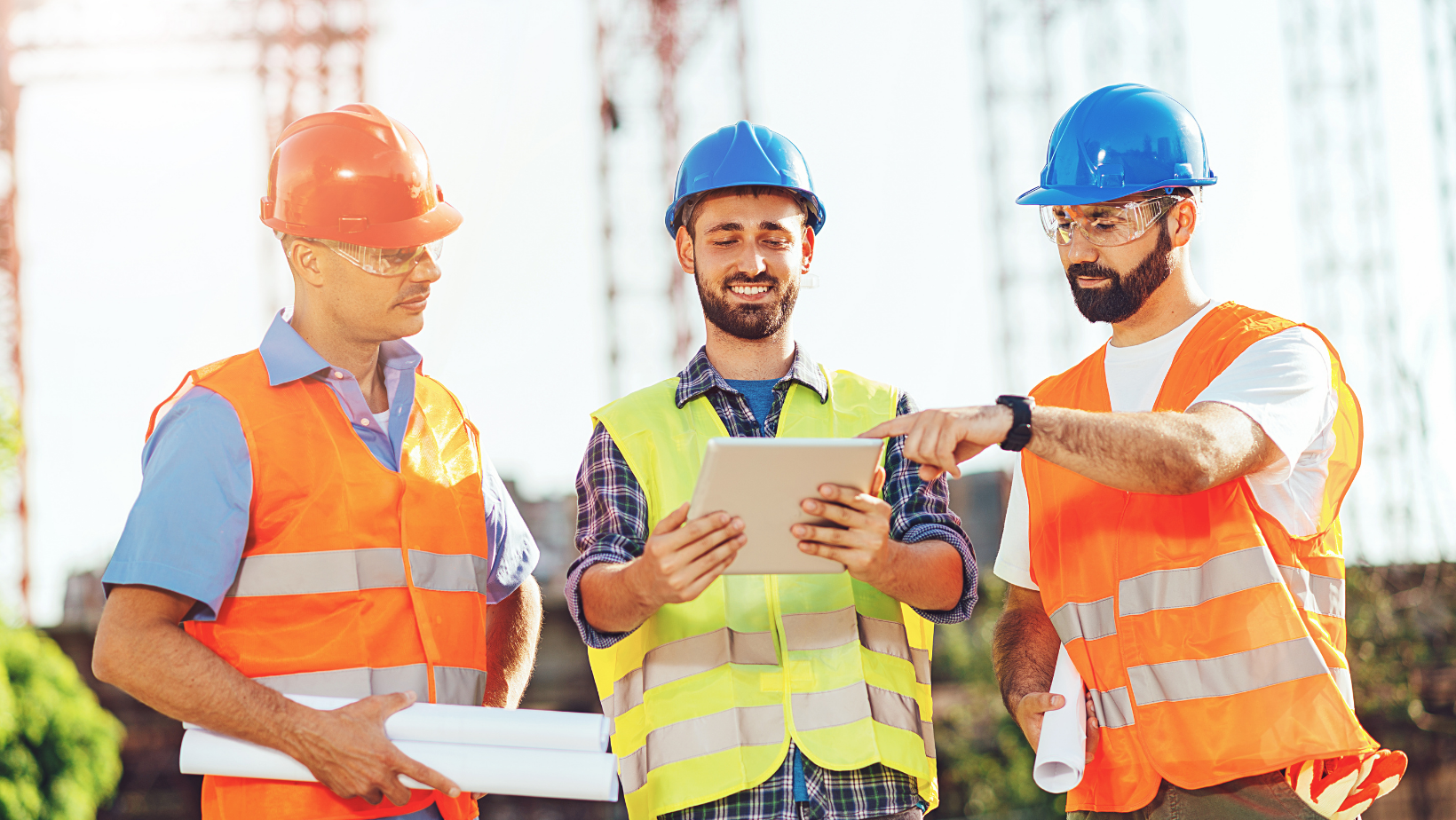 Three construction project managers with hard hats and reflective vests  on site with hardhats looking at an ipad or tablet.